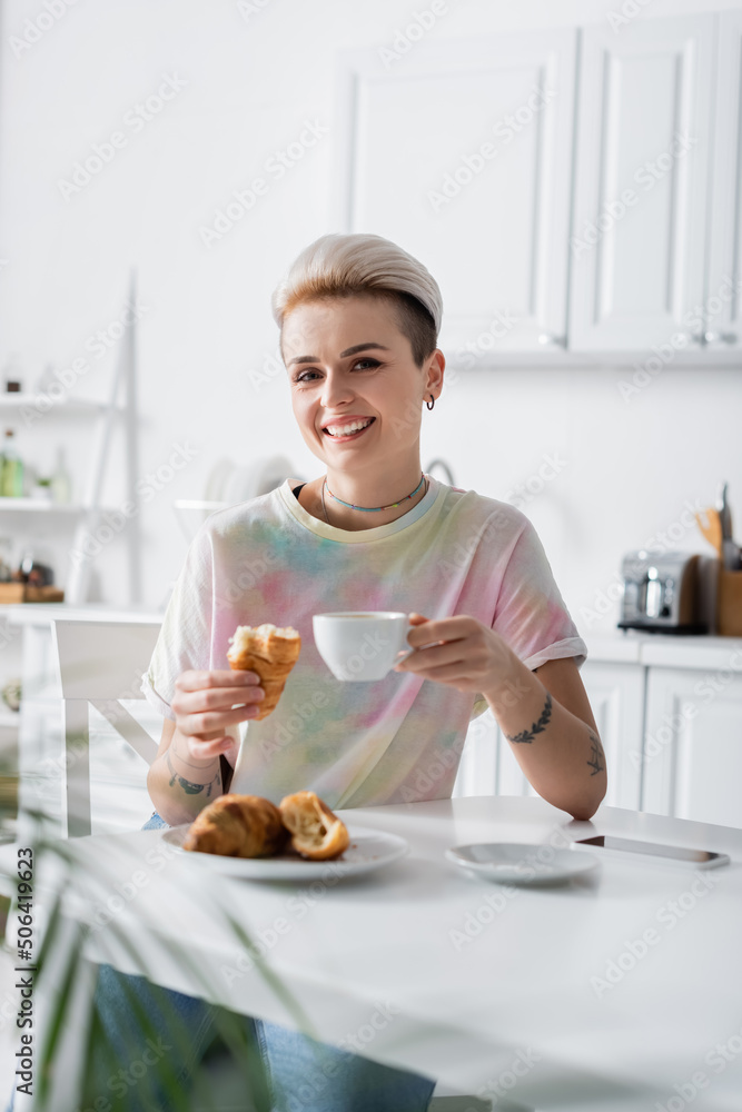 cheerful woman with coffee cup and tasty croissant looking at camera in kitchen cheerful woman with coffee cup and tasty croissant looking at camera in kitchen
