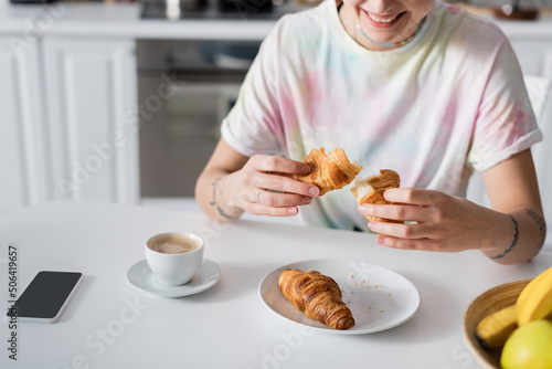 cropped view of smiling woman holding croissant near coffee cup and smartphone with blank screen