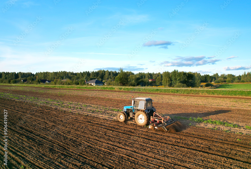 Obraz premium Tactor with disk harrow on plowing field. Cultivated land and soil tillage. Blue Tractor with disc cultivator on land cultivating. Agricultural tractor on soil cultivation field. Plough plowed.