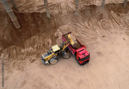 Wheel loader load sand in dump truck in open pit. Front end loader on earthworks in open-pit mining. Heavy machinery on developing opencast. Lorry truck sand transports from quarry, aerial view..