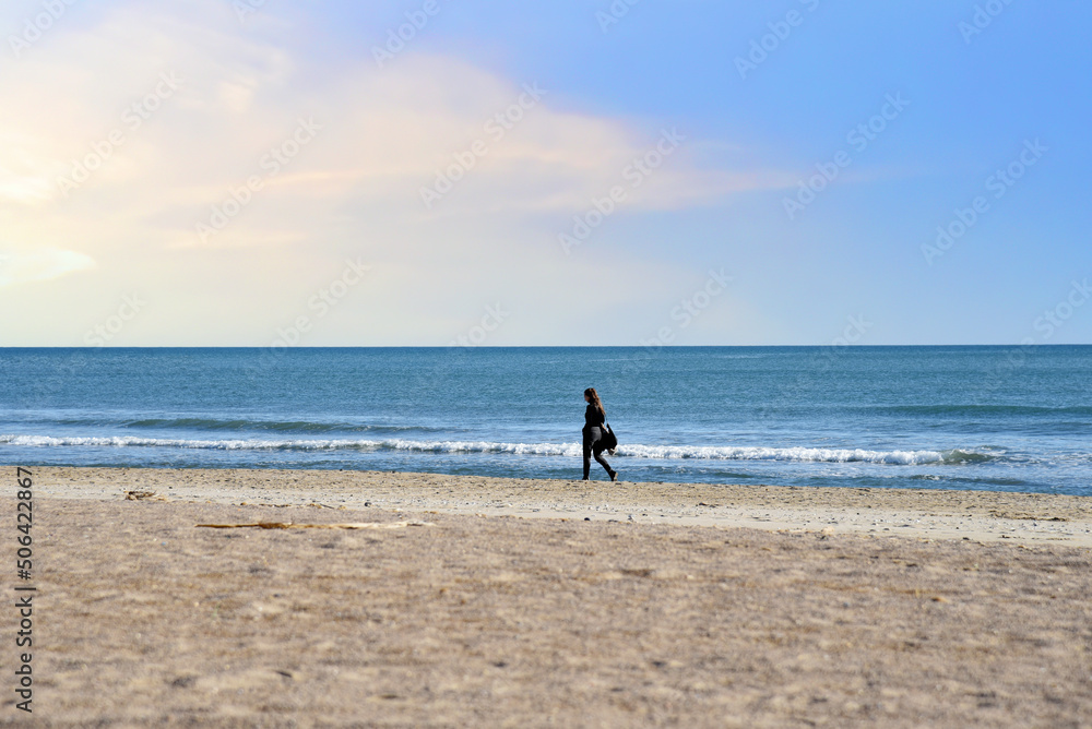 Girl is walking near the sea. Young woman walks along the sand of a beach near Mediterranean Sea against backdrop of waves and a stormy sky at sunset. Female person walks along coastline of the ocean.