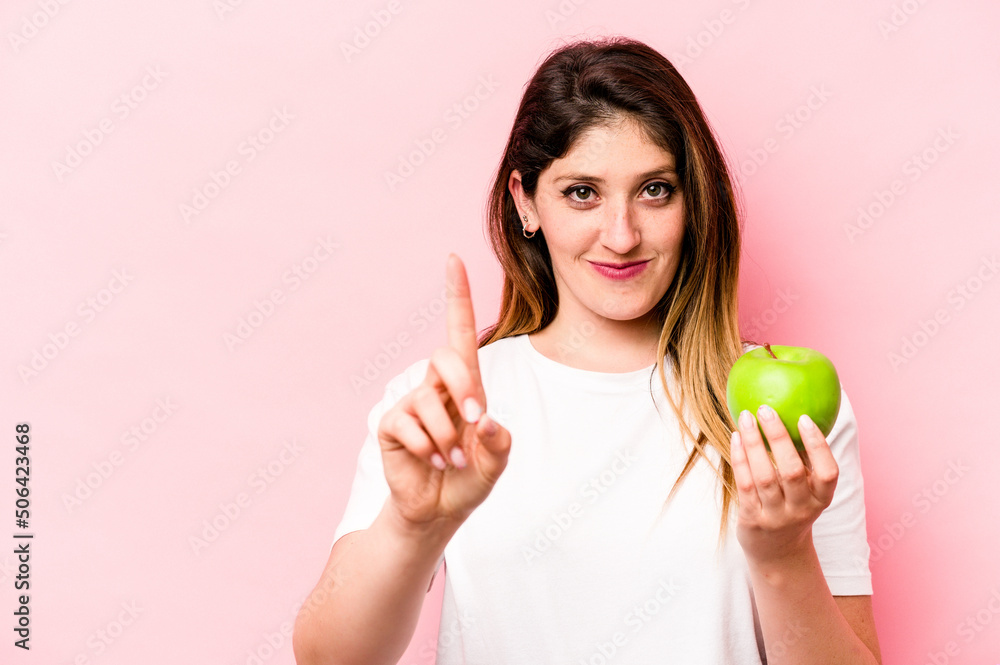 Young caucasian woman holding an apple isolated on pink background showing number one with finger. Young caucasian woman holding an apple isolated on pink background showing number one with finger.