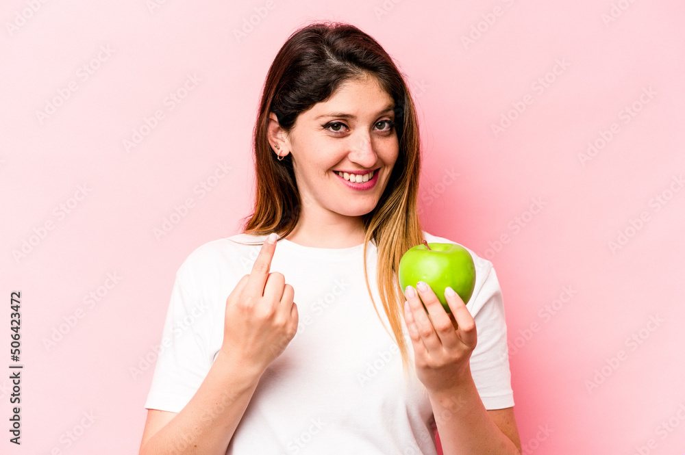 Young caucasian woman holding an apple isolated on pink background pointing with finger at you as if inviting come closer. Young caucasian woman holding an apple isolated on pink background pointing with finger at you as if inviting come closer.