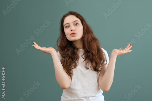 Teenage brunette girl in white t-shirt spreads her hands, on teal background. I don't know, confused gesture.