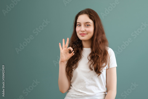 Teenage girl with blue eyes in white t-shirt is showing okay sign, smiling, on teal background.