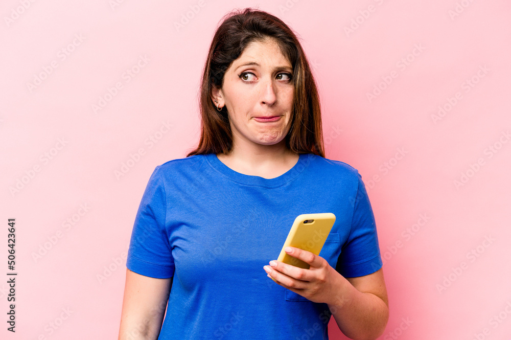 Young caucasian woman holding mobile phone isolated on pink background confused, feels doubtful and unsure. Young caucasian woman holding mobile phone isolated on pink background confused, feels doubtful and unsure.