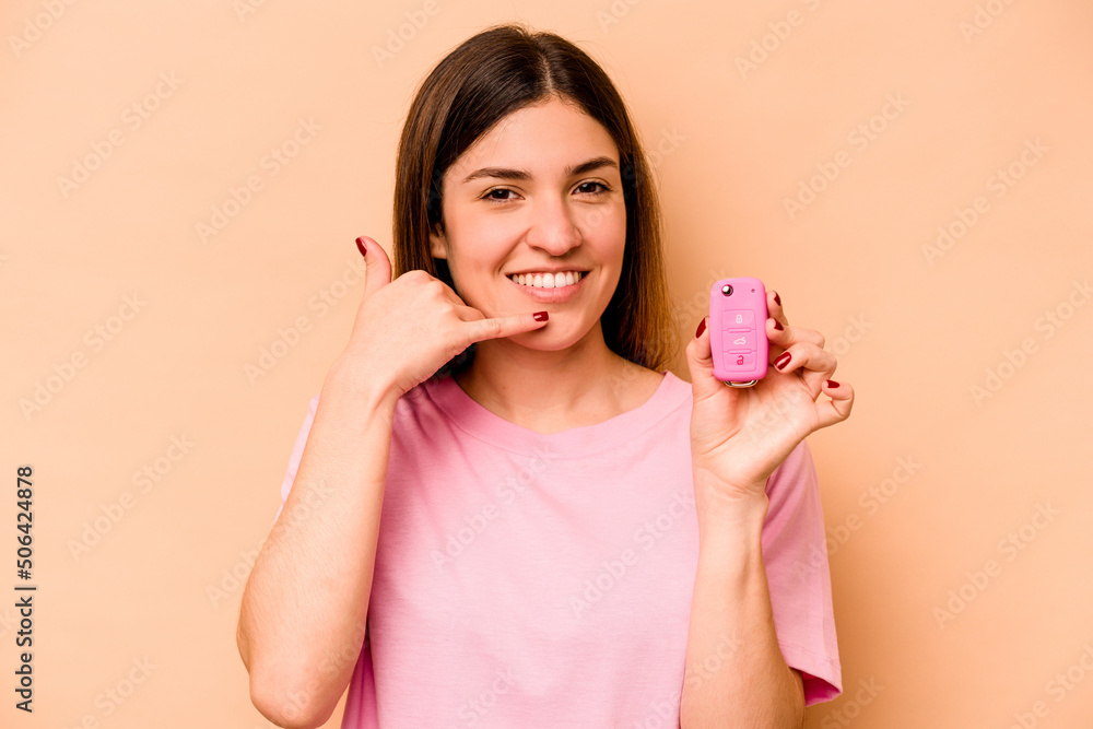 Young hispanic woman holding a keys car isolated on beige background showing a mobile phone call gesture with fingers. Young hispanic woman holding a keys car isolated on beige background showing a mobile phone call gesture with fingers.
