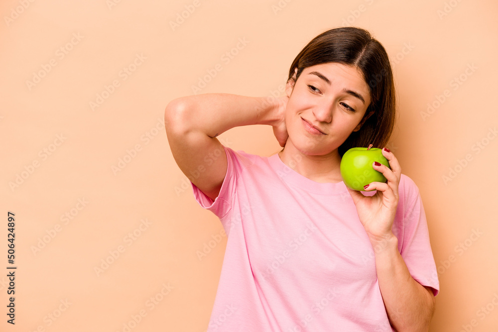 Young hispanic woman holding an apple isolated on beige background touching back of head, thinking and making a choice. Young hispanic woman holding an apple isolated on beige background touching back of head, thinking and making a choice.
