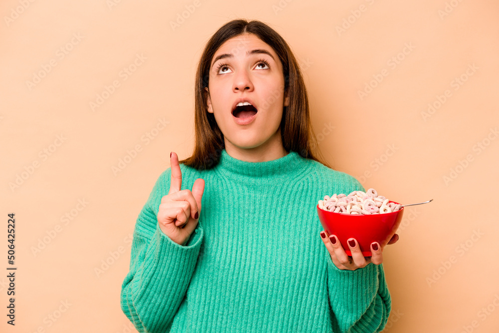 Young hispanic woman eating cereals isolated on beige background pointing upside with opened mouth. Young hispanic woman eating cereals isolated on beige background pointing upside with opened mouth.