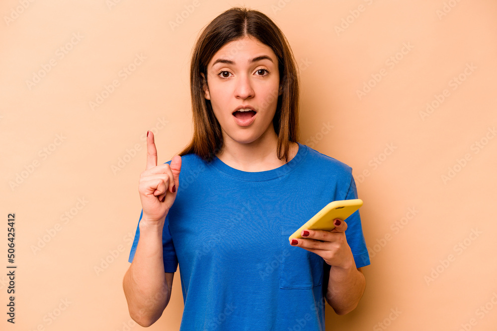 Young caucasian woman holding mobile phone isolated on beige background having an idea, inspiration concept. Young caucasian woman holding mobile phone isolated on beige background having an idea, inspiration concept.