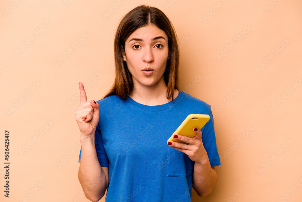 Young caucasian woman holding mobile phone isolated on beige background having some great idea, concept of creativity. Young caucasian woman holding mobile phone isolated on beige background having some great idea, concept of creativity.