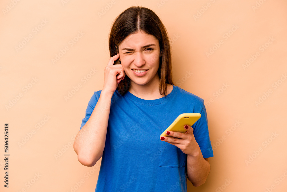 Young caucasian woman holding mobile phone isolated on beige background covering ears with hands. Young caucasian woman holding mobile phone isolated on beige background covering ears with hands.