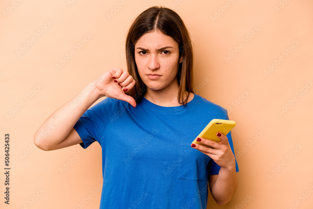 Young caucasian woman holding mobile phone isolated on beige background showing a dislike gesture, thumbs down. Disagreement concept. Young caucasian woman holding mobile phone isolated on beige background showing a dislike gesture, thumbs down. Disagreement concept.