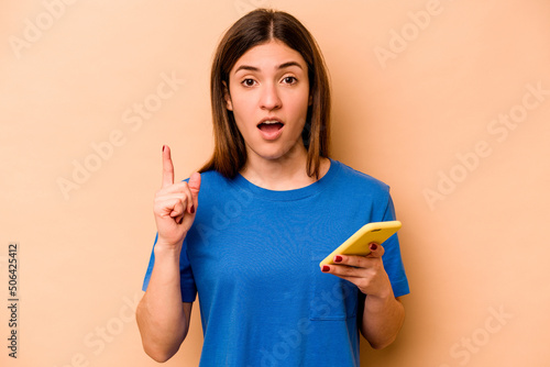 Young caucasian woman holding mobile phone isolated on beige background having an idea  inspiration concept.