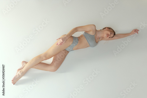 Full-length portrait of young slender woman in cotton underwear lying on floor, posing isolated over grey studio background