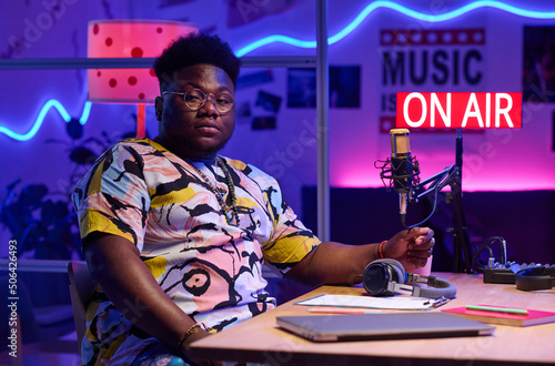 Horizontal medium portrait of confident young gen Z African American influencer wearing stylish casual clothes sitting at desk looking at camera