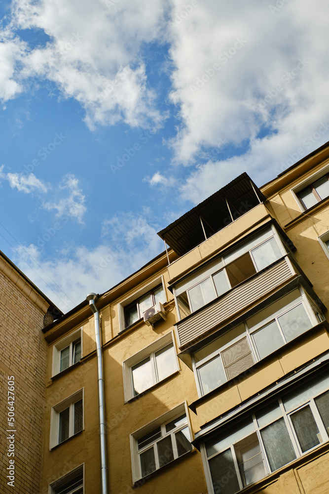 Low angle view of residential yellow house with balconies, windows and drainpipe. Blue sky with clouds. Air conditioner hangs on facade of house.