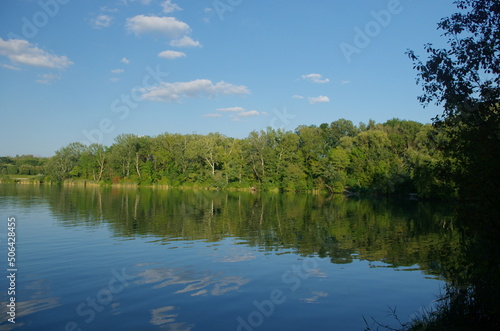 Natural panorama with lake and blue sky. Clouds in Lake Country. Summer panorama with green trees by the lake, beautiful and colorful reflections of trees, blue sky and clouds in the in the water. 