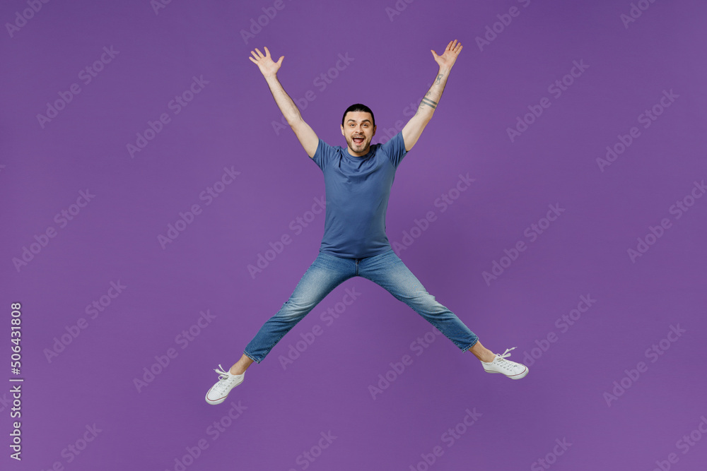 Full body young overjoyed excited cool man 20s wear basic blue t-shirt jump high with outstretched arms hands legs isolated on plain purple color background studio portrait. People lifestyle concept.