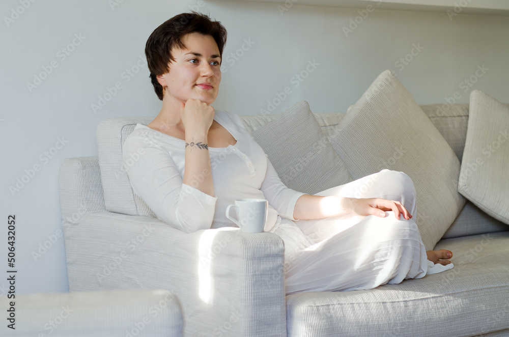 Lifestyle portrait of happy woman in white skirt, shirt relaxing, sitting on couch at cozy home. Drinking coffee. Dreaming Lifestyle portrait of happy woman in white skirt, shirt relaxing, sitting on couch at cozy home. Drinking coffee. Dreaming
