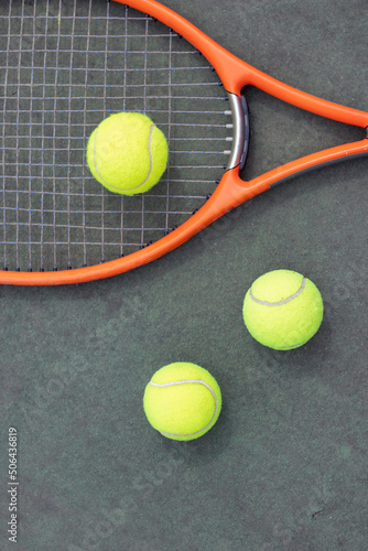 Nearby still life of tennis equipment consisting of an orange racket and three yellow balls of a green concrete court. © NOWRA photography