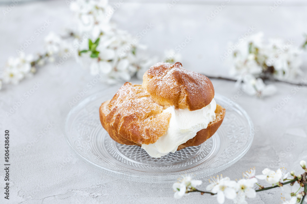 Choux Bun with whipped cream and sugar powder on top on glass plate. Gray background with spring white cherry blossom flowers. Delicate choux pastry dessert. French cream puff Choux Bun with whipped cream and sugar powder on top on glass plate. Gray background with spring white cherry blossom flowers. Delicate choux pastry dessert. French cream puff