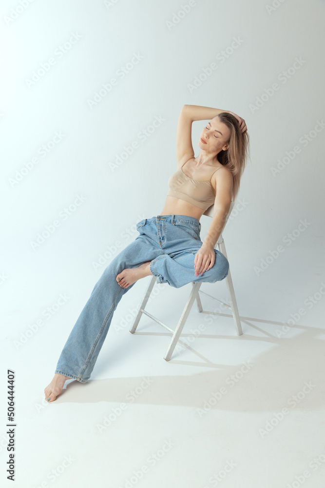 Portrait of tender young girl sitting on chair, posing in beige underwear and big jeans isolated over grey studio background. Self-love
