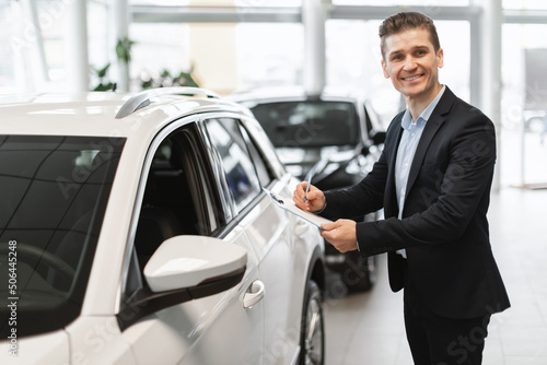 Happy young auto insurance agent standing near new car, checking it, taking notes, working at automobile dealership © Prostock-studio