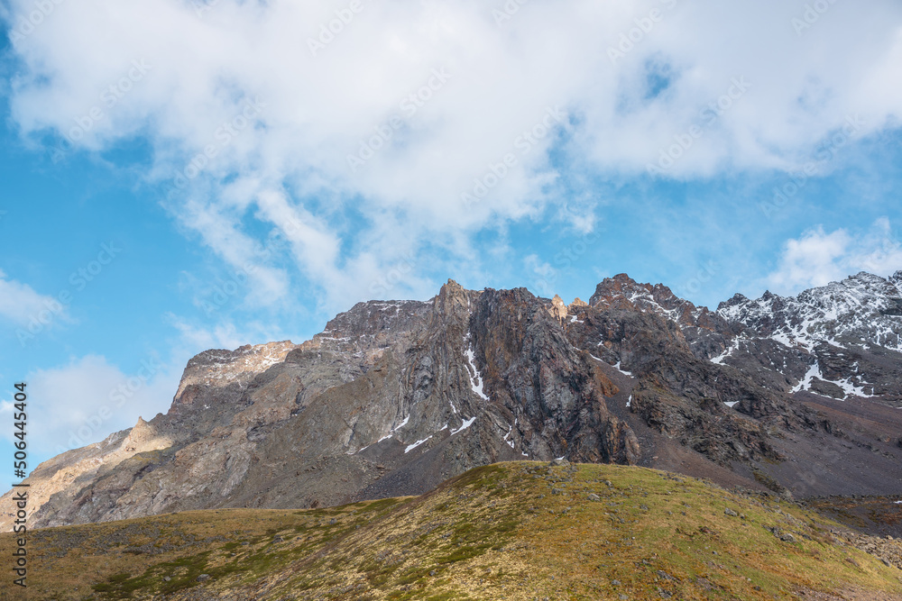 Atmospheric landscape with sharp pinnacle on sunlit high mountain range under clouds in blue sky. Beautiful alpine scenery with peaked mountain top in sunlight under cloudy sky at changeable weather. Atmospheric landscape with sharp pinnacle on sunlit high mountain range under clouds in blue sky. Beautiful alpine scenery with peaked mountain top in sunlight under cloudy sky at changeable weather.
