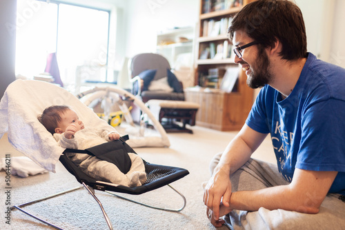 Father interacting with newborn baby in bouncer with messy house in background photo