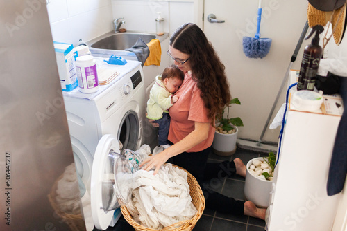 Young mum taking wet clean clothes out of washing machine while holding newborn baby photo