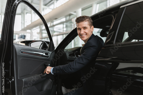 Successful millennial car salesman sitting inside auto salon, looking at camera through open door in modern dealership © Prostock-studio