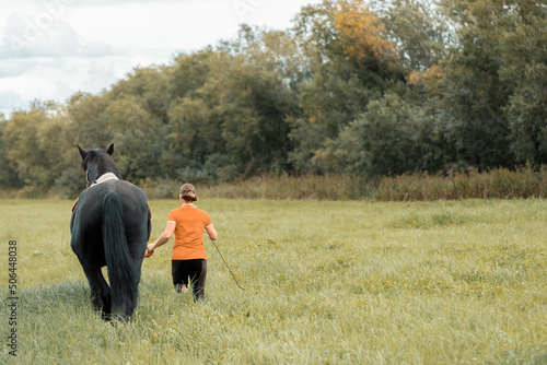 Rider leading black horse on grass, rear view. © Ирина Орлова