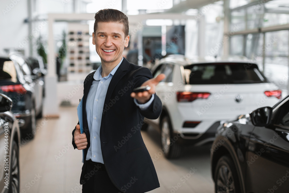 Successful young salesman giving car key at camera in modern dealership, empty space. Vehicle local distribution concept