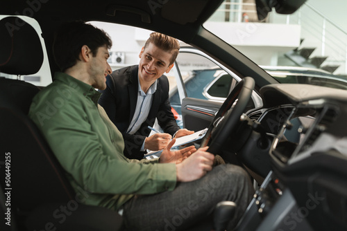 Young Caucasian guy clarifying car purchase details with salesman, sitting inside auto salon at dealership center