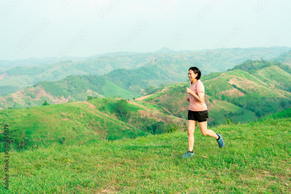 Obraz premium Young Asian woman runner, wearing black sportswear, running on a big mountain trail, cool morning, windmills, and sky in the background.