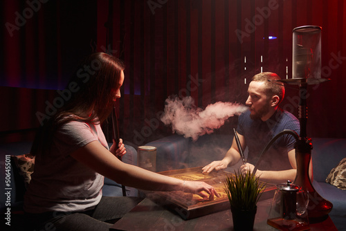 a young man and a woman emit white hookah smoke from their mouths in a relaxed atmosphere and play backgammon, smoke a hookah in a dark hookah room
