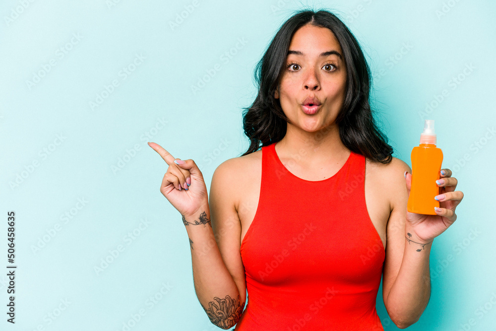 Young hispanic woman holding sun cream isolated on blue background pointing to the side Young hispanic woman holding sun cream isolated on blue background pointing to the side
