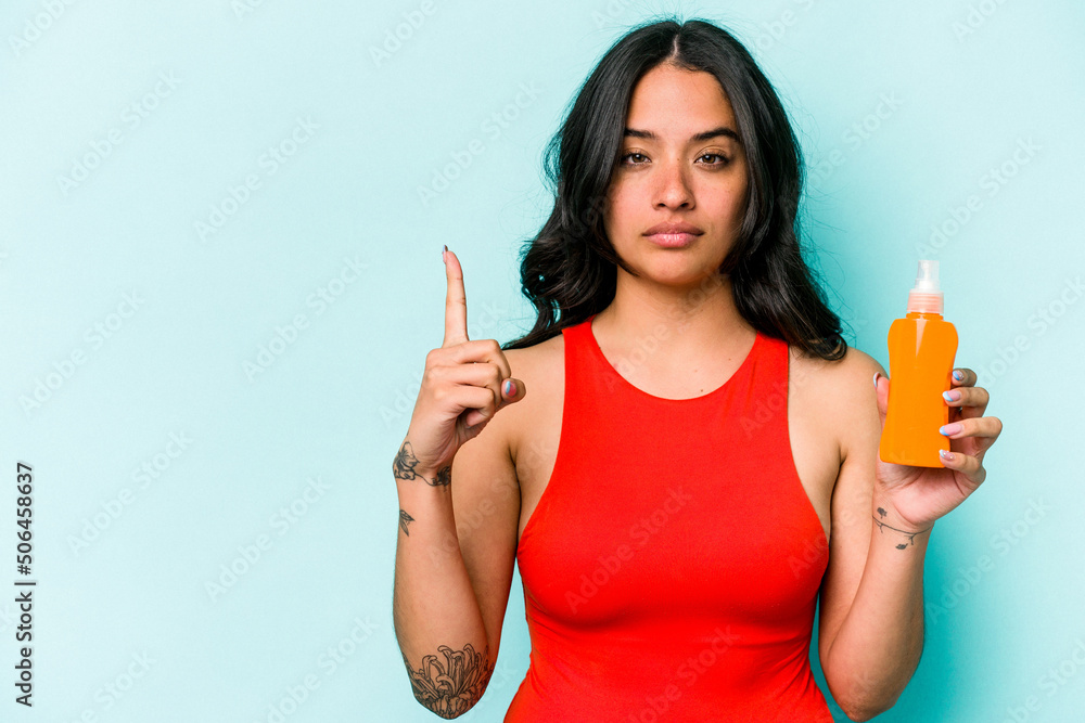 Young hispanic woman holding sun cream isolated on blue background showing number one with finger. Young hispanic woman holding sun cream isolated on blue background showing number one with finger.