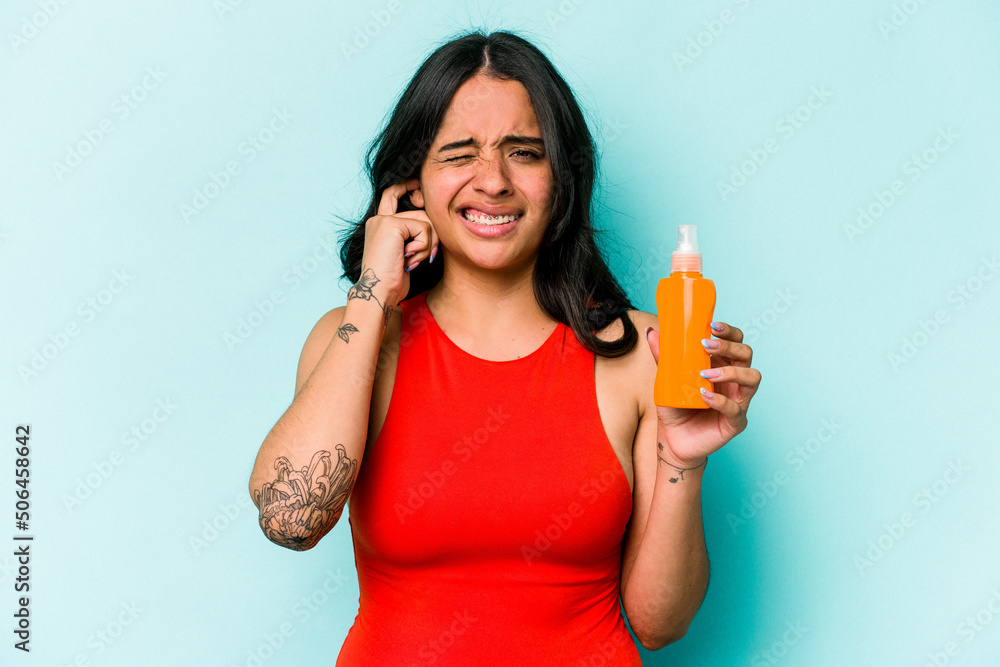 Young hispanic woman holding sun cream isolated on blue background covering ears with hands. Young hispanic woman holding sun cream isolated on blue background covering ears with hands.