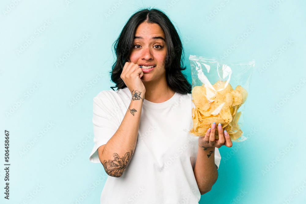 Young hispanic woman holding a bag of chips isolated on blue background biting fingernails, nervous and very anxious. Young hispanic woman holding a bag of chips isolated on blue background biting fingernails, nervous and very anxious.