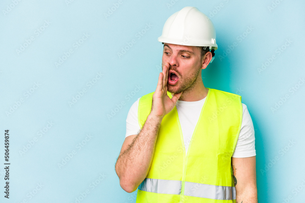 Young laborer caucasian man isolated on blue background is saying a secret hot braking news and looking aside