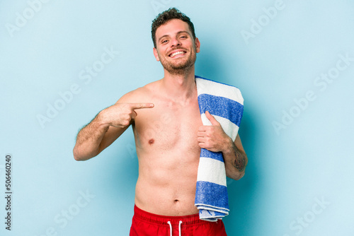 Young caucasian man holding beach towel isolated on blue background person pointing by hand to a shirt copy space, proud and confident
