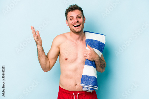 Young caucasian man holding beach towel isolated on blue background receiving a pleasant surprise, excited and raising hands.