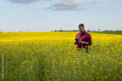 Middle age farmer standing in rapeseed field with digital tablet examining crop.