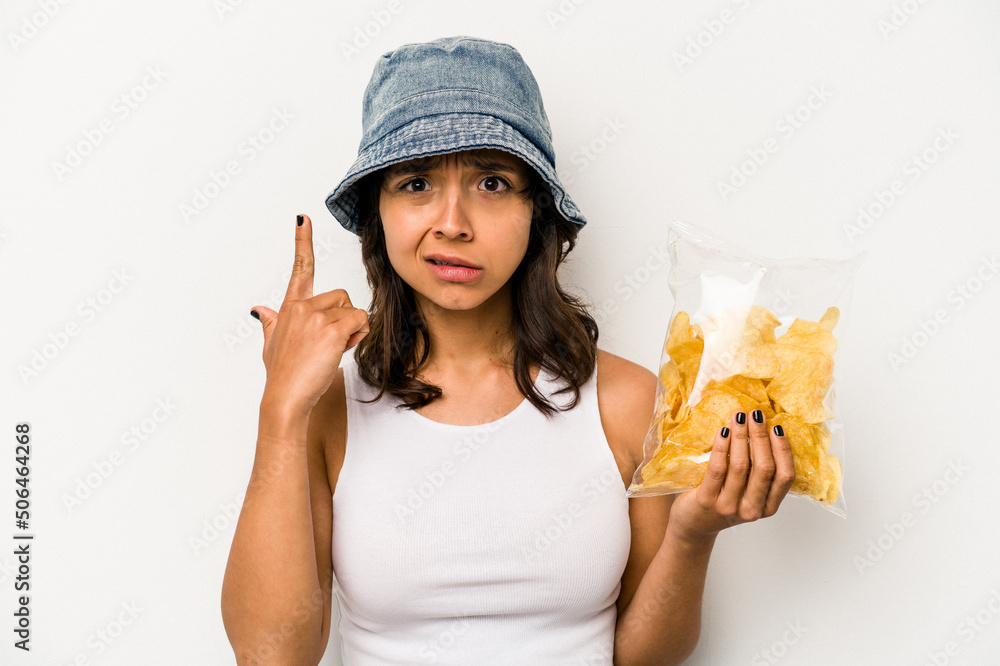 Young hispanic woman holding a bag of chips isolated on white background showing a disappointment gesture with forefinger. Young hispanic woman holding a bag of chips isolated on white background showing a disappointment gesture with forefinger.