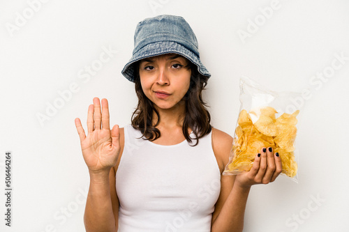 Young hispanic woman holding a bag of chips isolated on white background standing with outstretched hand showing stop sign  preventing you.