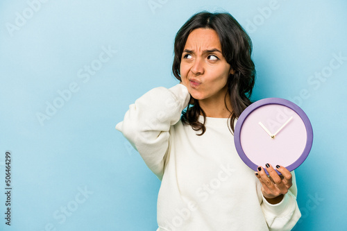 Young hispanic woman holding a clock isolated on blue background touching back of head, thinking and making a choice. photo