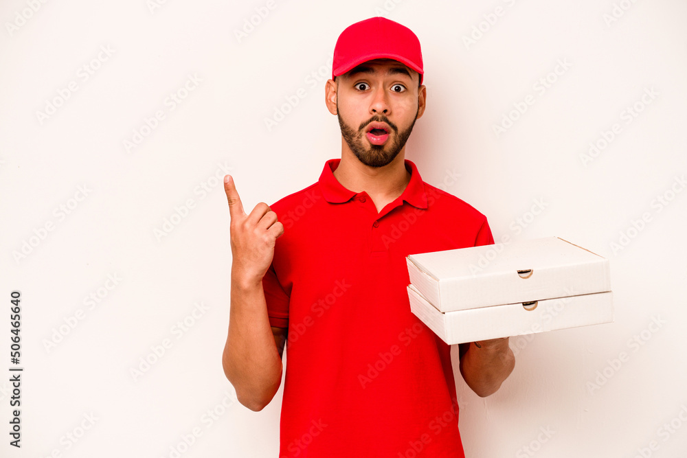 Young hispanic delivery man holding pizzas isolated on white background pointing to the side Young hispanic delivery man holding pizzas isolated on white background pointing to the side