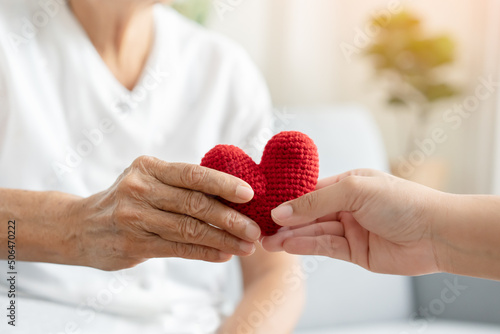 Young and senior woman holding each other hands and red yarn heart shape togetherness concept. Elderly care and protection with love from grandchild.